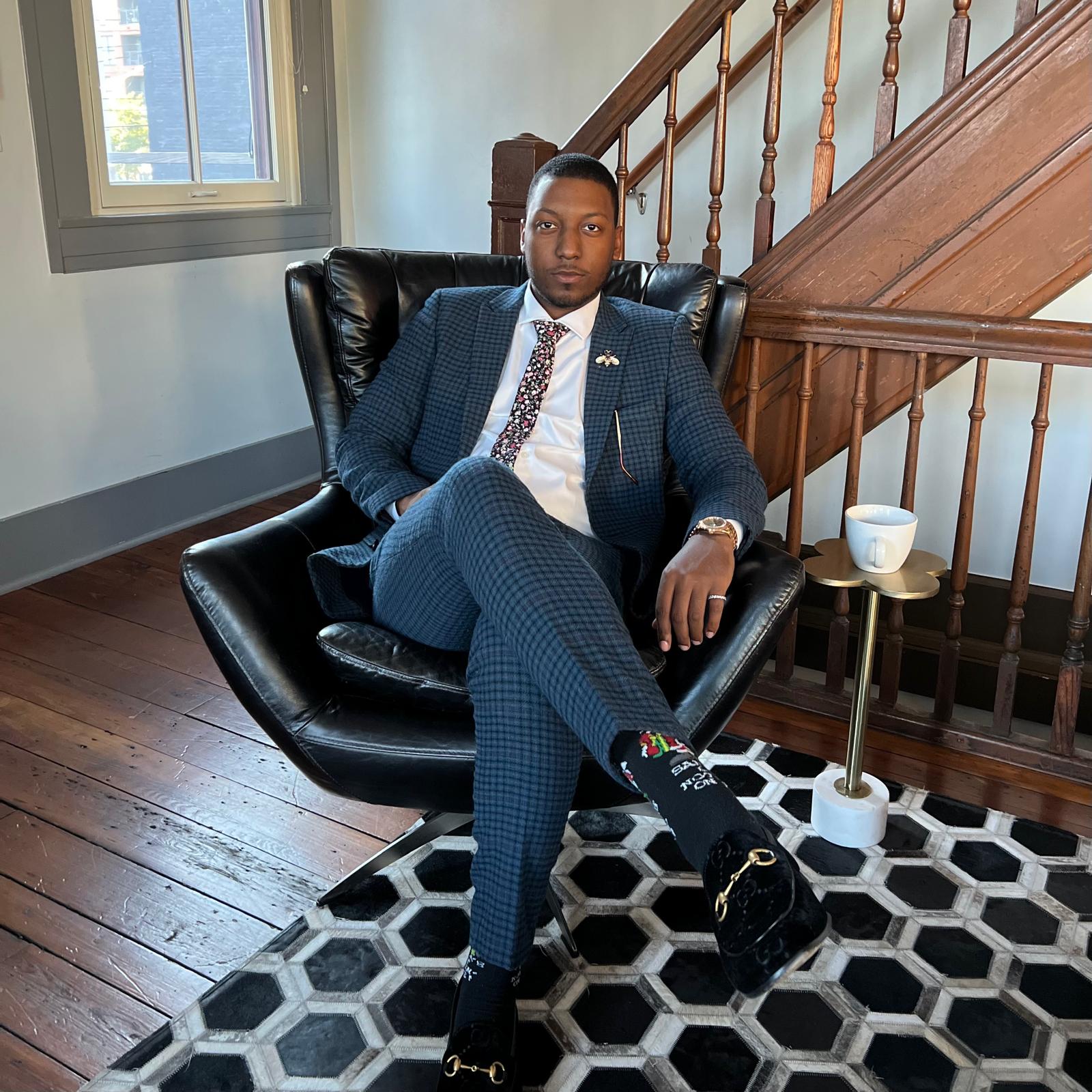 Aton Williams seated in an armchair beside a wooden staircase